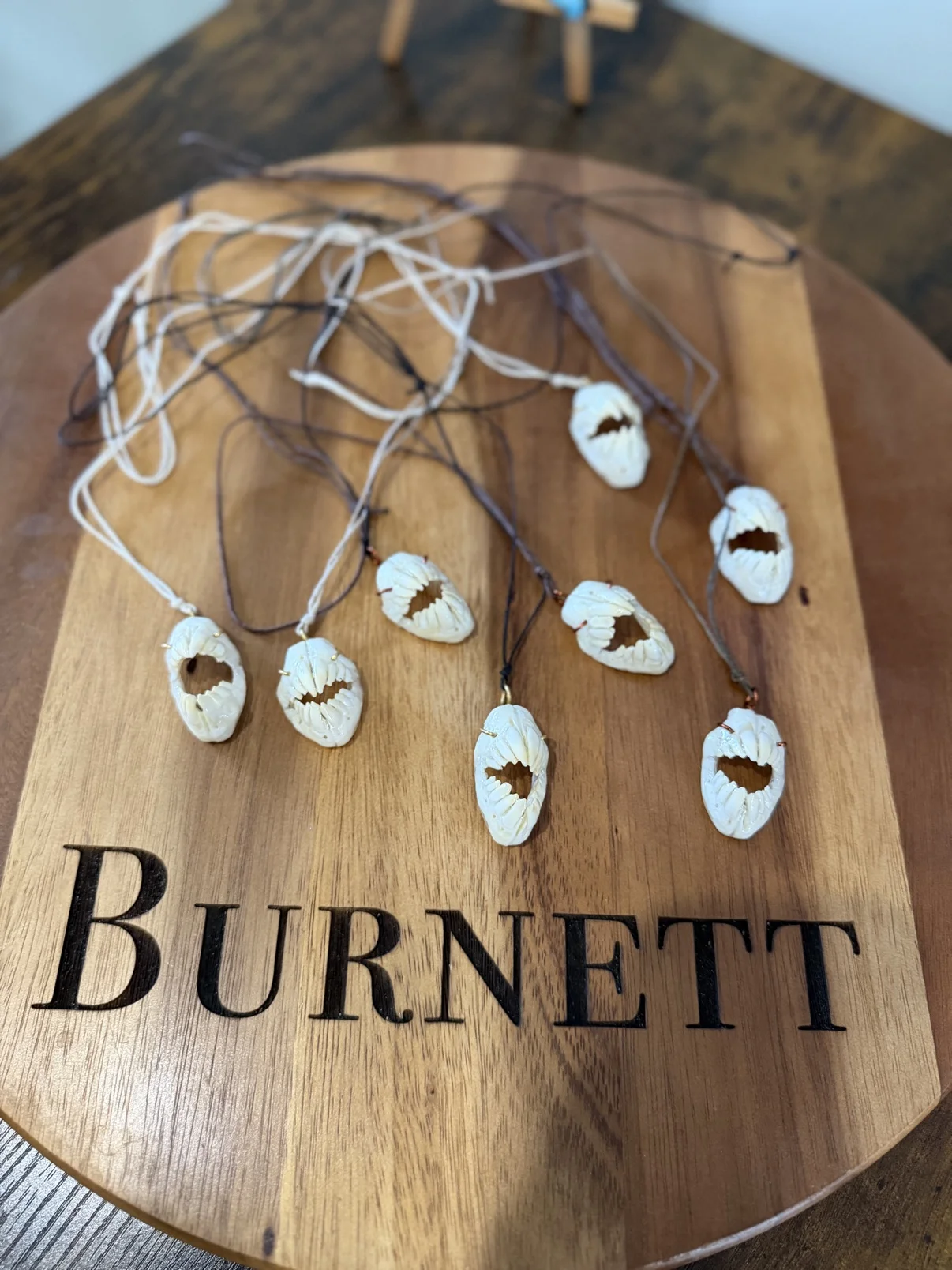 Photograph of seven of my triggerfish jaw necklaces laid out on a round wooden board engraved with the name Burnett, white bone jaw pendants on white, brown and black cords