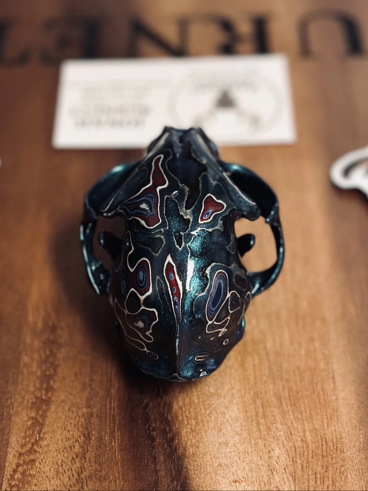 Photograph of my small hand-painted animal skull from above on a wooden board, showing the red and white fordite patterning across the dark teal top of the skull