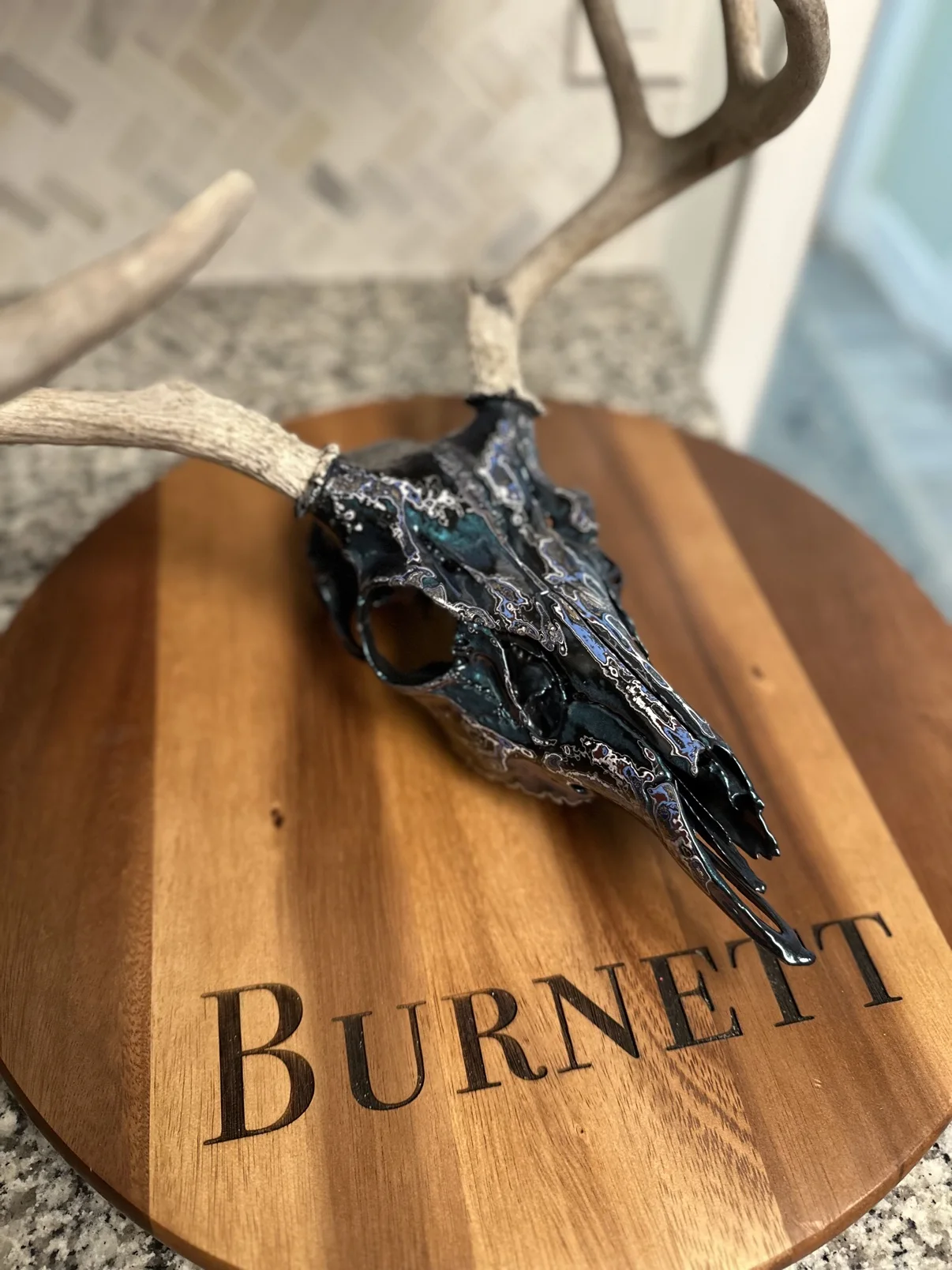Photograph of my hand-painted whitetail deer skull from an angled view on a round wooden board, black finish with teal, blue and silver fordite marbling running down the snout