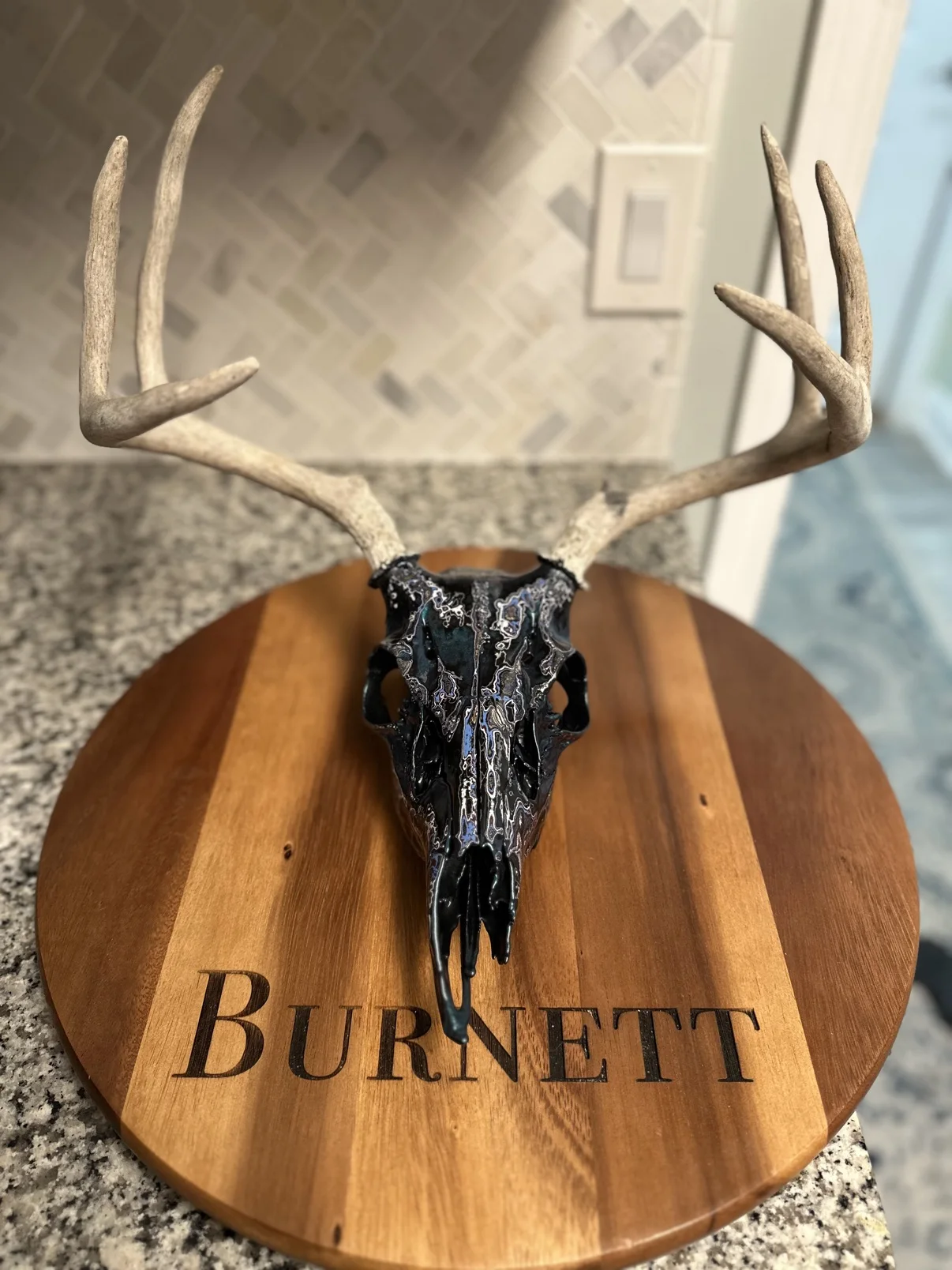 Photograph of my hand-painted whitetail deer skull from the front on a round wooden board, black finish with teal, blue and silver fordite marbling and natural antlers