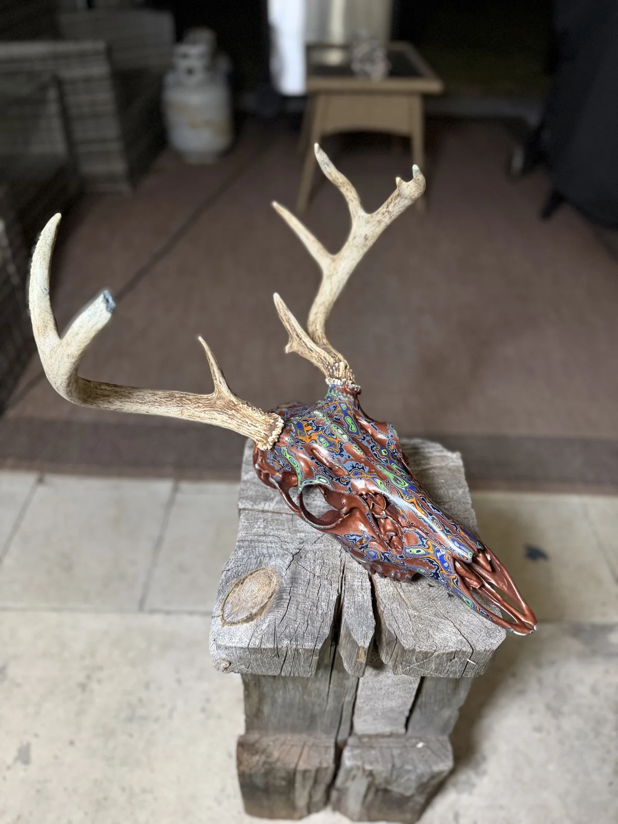 Photograph of my hand-painted whitetail deer skull from an angled view on a wooden post, copper and brown finish with bright multicolor fordite patterning running down the snout