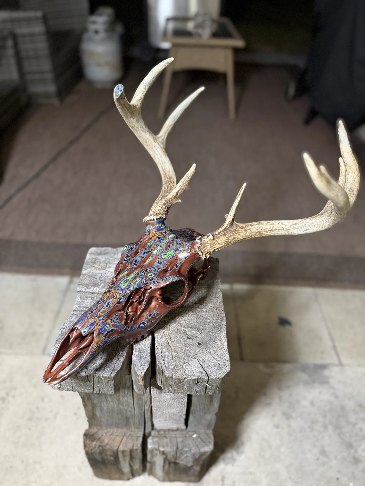 Photograph of my hand-painted whitetail deer skull from the side on a wooden post, showing the profile of the copper skull with multicolor fordite patterning and the natural antlers
